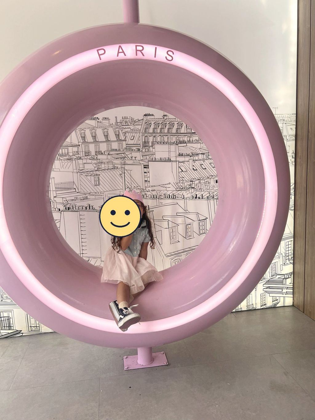 A child in a paper crown sitting inside a giant pink PARIS ring against a backdrop of Paris rooftops