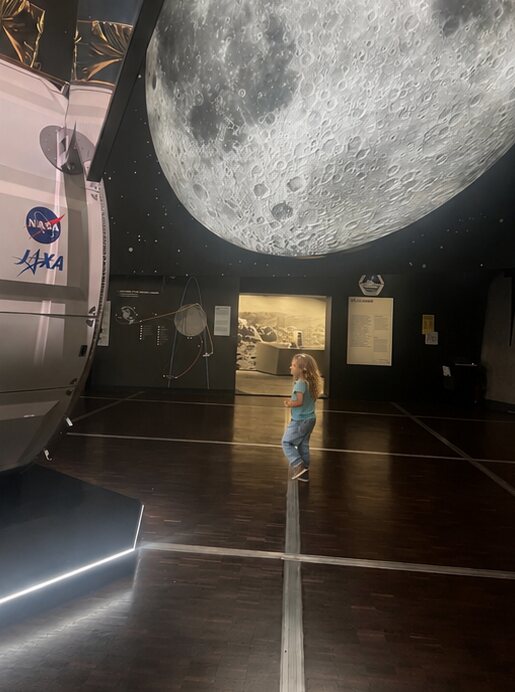 A child standing under a giant illuminated moon beside a NASA space capsule at the Cité des Sciences, Paris